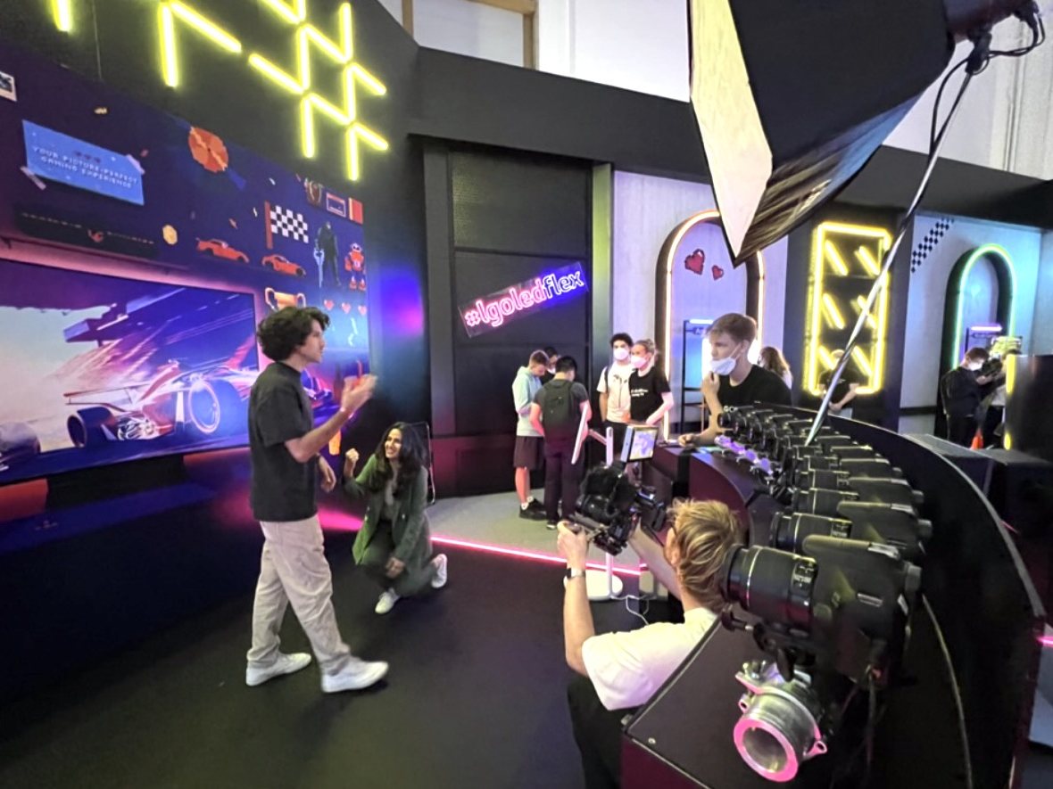 A curved row of cameras and an operator face two guests posing in front of a pixel-art wall and a neon sign